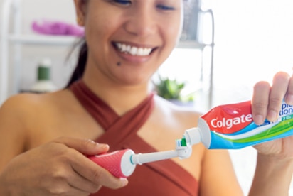 En kvinde, der smiler, mens hun lægger Colgate-tandpasta på en tandbørste på et badeværelse.